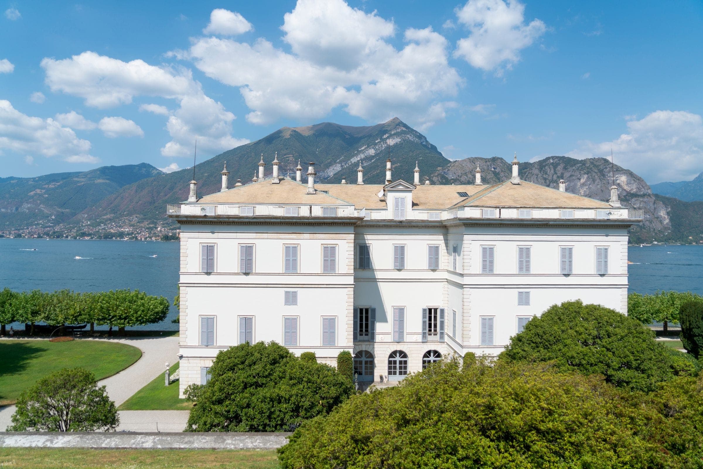 A white lakefront villa and its gardens below wooded mountains
