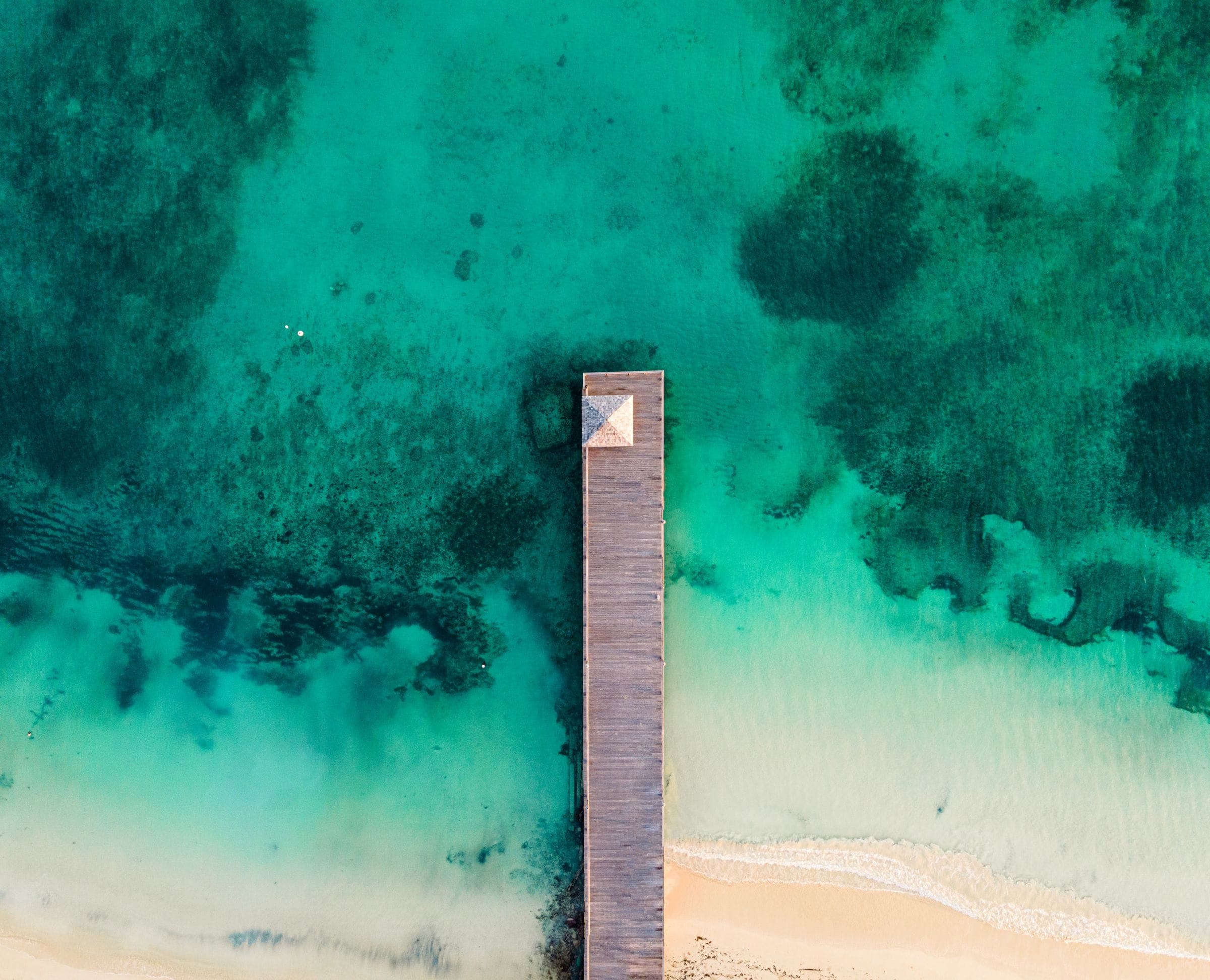 A jetty reaching over pale turquoise shallows, photographed from above